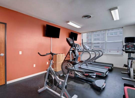 A gym at Latitude Apartments with a stationary bike, treadmills, and an elliptical facing two wall-mounted TVs. Large windows let in natural light, creating a clean, energizing atmosphere.