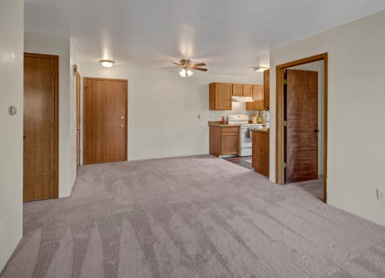 Spacious living area here at Legacy West Apartments with beige carpet and neutral walls, connected to a kitchen with wooden cabinets and white appliances. Ceiling fan above adds warmth.