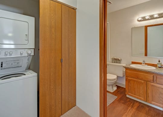 Laundry area here at Legacy West Apartments with a stacked washer and dryer next to wooden cabinets. Adjacent bathroom with sink, mirror, toilet, and warm lighting; cozy atmosphere.