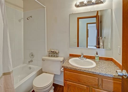 Small bathroom here at Legacy West Apartments with white tiled shower-tub combo, beige curtain, wooden vanity, granite counter, oval sink, mirror, and chrome light fixture.