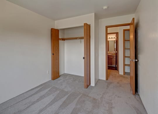Empty bedroom here at Legacy West Apartments with light gray carpet, open wooden closet doors, and an adjoining bathroom door. Neutral walls and a tidy ambiance.
