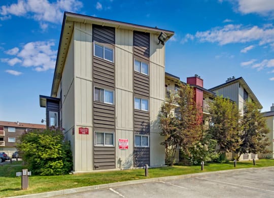 Three-story Legacy West apartment building with tan and brown siding, surrounded by trees and grass. Clear blue sky, empty parking lot in the foreground.