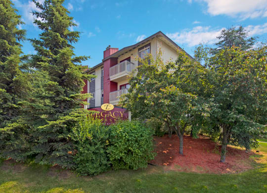 Legacy West Apartment building surrounded by lush trees and greenery under a blue sky. A sign reads "Legacy" in the foreground, creating a peaceful ambiance.