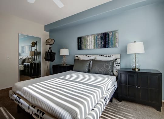 Modern bedroom here at Rio Paradiso Apartments with a neatly made bed featuring patterned pillows. Soft blue walls are adorned with abstract art. Sunlight casts striped shadows, creating a calm ambiance.
