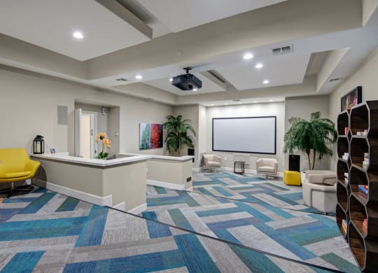 Modern meeting room here at Rio Paradiso Apartments with patterned blue carpet, cream walls, and a large central screen. Comfortable seating, potted plants, and a vibrant, open feel.