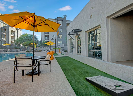 Modern outdoor courtyard here at Rio Paradiso Apartments with yellow umbrellas, tables, and chairs by a pool. A concrete building and grassy area with a cornhole game create a relaxing atmosphere.