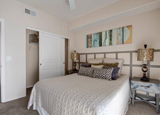 A cozy bedroom here at Rio Paradiso Apartments with a neatly made bed featuring patterned pillows. The headboard has a geometric design, flanked by two lamps. Abstract art hangs above, creating a serene and stylish ambiance.