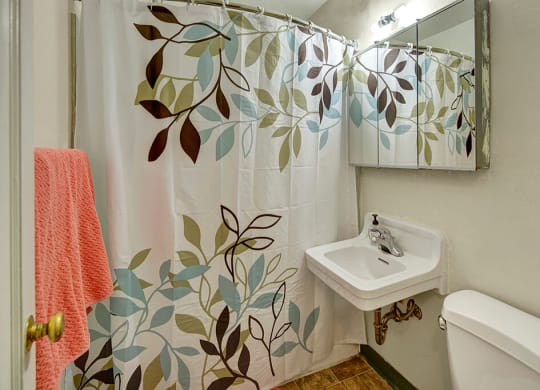 Bathroom here at Mendenhall Tower Apartements with a leafy pattern shower curtain, white sink, and mirror. A coral towel hangs nearby, creating a calm, fresh atmosphere.