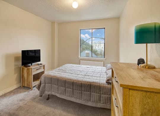 A cozy bedroom here at Mendenhall Tower Apartment featuring a wooden bed with a gray quilt, a small TV on a stand, a dresser with a teal lamp, and a window with a view of trees.
