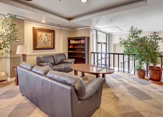 Cozy lounge area here at Mendenhall Tower Apartments with two gray leather sofas facing a round, wooden coffee table. A bookshelf, framed artwork, and large potted plants enhance the warm atmosphere.