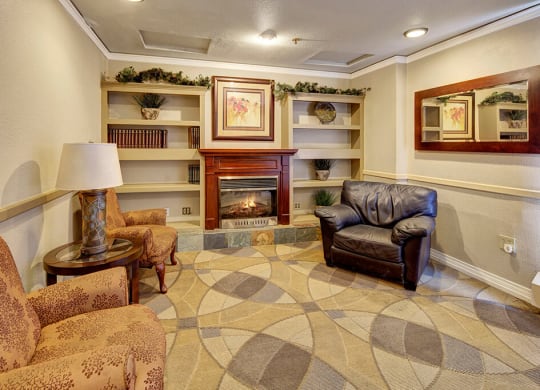 Cozy lounge area here at Mendenhall Tower Apartments with a fireplace, flanked by a brown leather chair and floral-patterned sofas. Bookshelves, a framed painting, and a mirror enhance warmth.