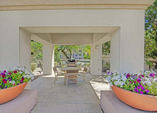Outdoor patio here at Mirador Apartments with covered seating around a table and a grill in the background. Colorful flower pots on each side. Sunny and tranquil atmosphere.
