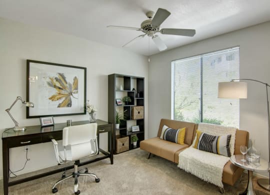 Modern home office here at Mirador Apartments with a white chair, dark desk, and a leaf art piece on the wall. A tan sofa with striped pillows sits by a large window.