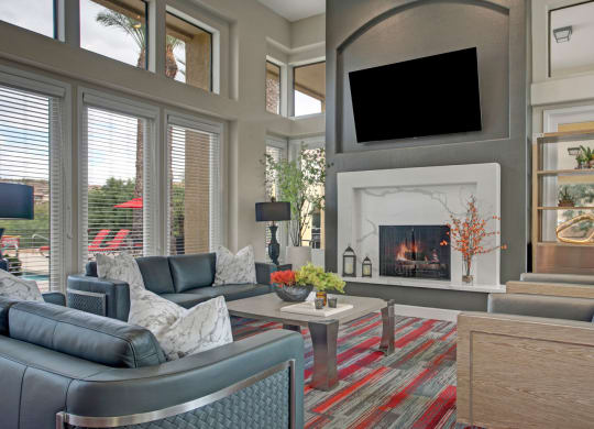 Modern resident lounge here at Mirador Apartments with large windows, gray sofas, a red-patterned rug, and a fireplace. A TV is mounted above the mantle. Room feels cozy and inviting.