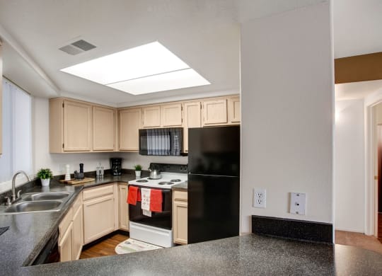 Bright kitchen Mirador Apartments with light wood cabinets, black appliances, and dark granite countertops. Decor includes potted plants and red towels. Cozy and modern feel.