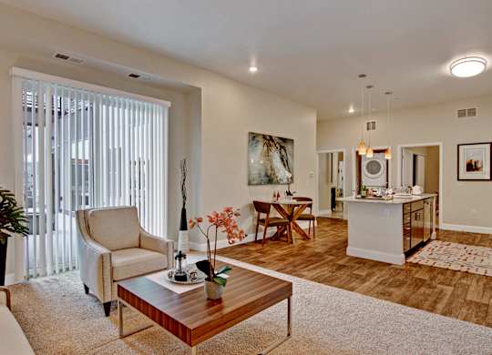 A welcoming open-concept living area here at 4800 Excelsior featuring a comfortable seating arrangement with a lounge chair and coffee table set on soft carpeting beside large vertical windows. The space flows into a dining nook and modern kitchen with an island, pendant lighting, and warm wood-style flooring, creating a bright and inviting home layout.