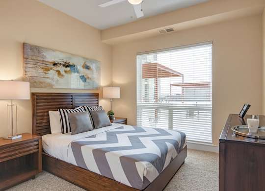 A welcoming bedroom here at 4800 Excelsior featuring a wood bed frame with patterned bedding and coordinated accent pillows. Matching nightstands with table lamps sit beneath a large abstract art piece, while plush carpeting, neutral walls, and a wide window with blinds bring in natural light and create a calm, comfortable retreat.