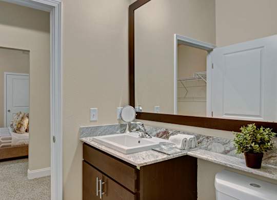 A stylish bathroom here at 4800 Excelsior featuring a stone countertop vanity with an undermount sink, warm wood cabinetry, and a large framed mirror. Open shelving holds neatly folded towels, while neutral walls and clean finishes create a bright, polished space with a connected view toward the bedroom and closet area.