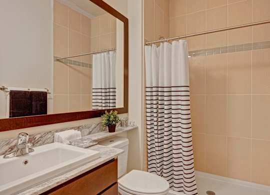 A clean bathroom here at 4800 Excelsior featuring a stone countertop vanity with an undermount sink, warm wood cabinetry, and a large framed mirror. A tiled tub and shower with a patterned curtain sits beside the vanity, while neutral tile walls, soft lighting, and neatly placed towels create a calm, well-organized space.