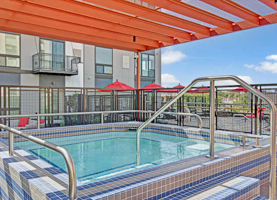 A modern outdoor spa here at 4800 Excelsior featuring a tiled hot tub with built-in steps and stainless handrails beneath a bold orange pergola. The fenced courtyard includes contemporary apartment façades, red patio umbrellas, and seating areas, creating a vibrant, inviting space for relaxation and socializing.