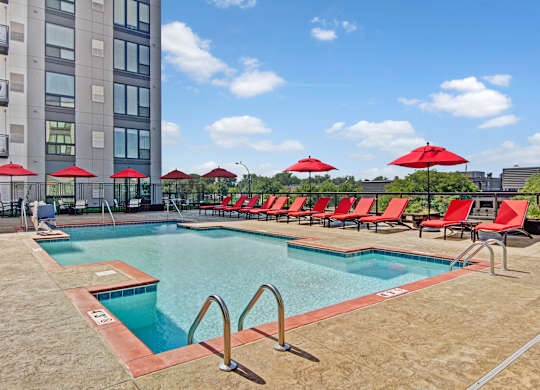 A resort-style outdoor pool here at 4800 Excelsior showcasing a rectangular swimming pool with clear blue water, stainless handrails, and a spacious concrete deck. Red lounge chairs and matching umbrellas line the poolside, with modern apartment buildings and treetop views creating an open, relaxing atmosphere.