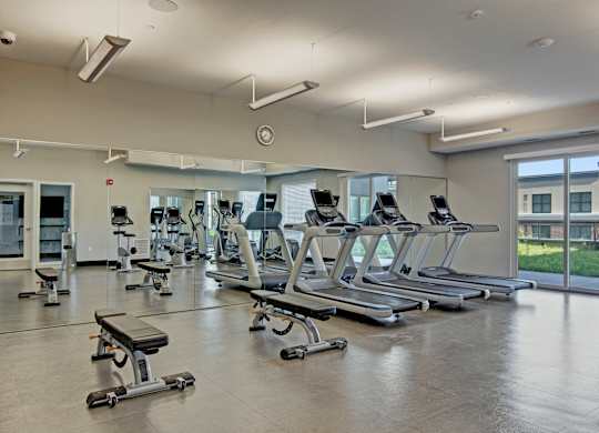A modern fitness center here at 4800 Excelsior featuring multiple treadmills, weight benches, and cardio equipment arranged across a spacious open floor. Large wall mirrors and overhead lighting enhance the bright atmosphere, while tall windows bring in natural light and views of the surrounding community.