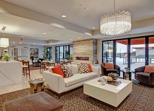 A stylish resident lounge here at 4800 Excelsior featuring a modern seating area with a white sofa, accent chairs, and decorative tables arranged around a contemporary fireplace. The space includes a large chandelier, a sleek bar with counter seating, and floor-to-ceiling windows that open to an outdoor terrace, creating an inviting area for relaxing or gathering.