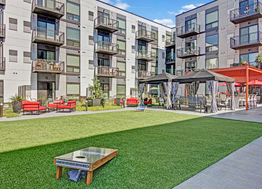 A landscaped courtyard here at 4800 Excelsior featuring a wide green lawn with a cornhole game, surrounded by modern apartment buildings with private balconies. Outdoor lounge seating with red cushions, shaded cabanas, and a nearby pergola create a relaxed, social setting designed for gathering, recreation, and everyday outdoor enjoyment.