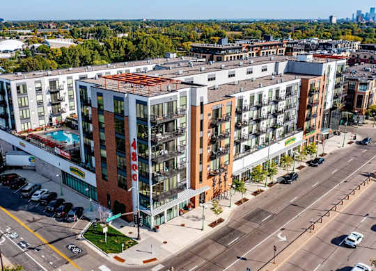 Aerial exterior view here at 4800 Excelsior showcasing a modern mid-rise apartment community with brick and light-toned façades, private balconies, and rooftop amenity spaces. The building sits along a city street with retail at ground level, surrounding greenery, nearby neighborhoods, and expansive treetop views extending toward the skyline.
