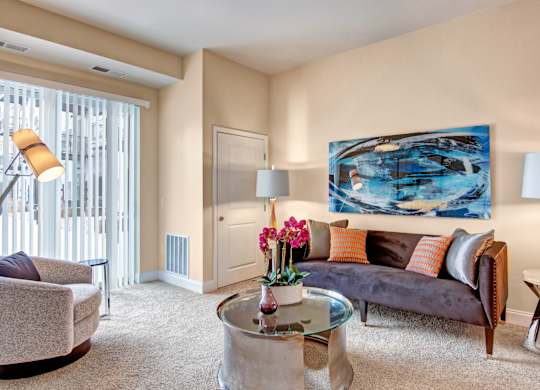A cozy living room here at 4800 Excelsior featuring plush carpeting, a contemporary sofa with accent pillows, and a round glass coffee table centered in the space. Soft lighting, abstract wall art, and a large window with vertical blinds create a warm, comfortable atmosphere with plenty of natural light.