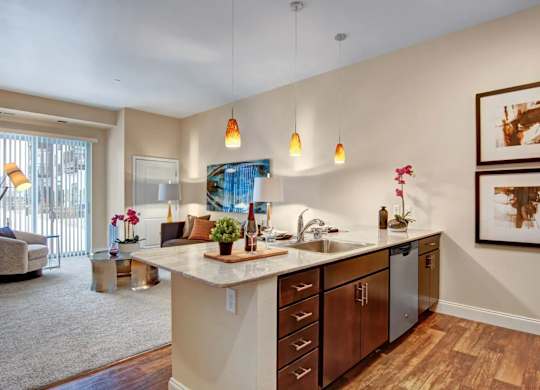 A contemporary kitchen here at 4800 Excelsior showcasing a spacious peninsula with light countertops, an undermount sink, and dark wood cabinetry accented by sleek hardware. Pendant lights hang above the counter, while stainless steel appliances and warm wood-style flooring connect the kitchen to the adjacent living area with soft carpeting and modern décor.