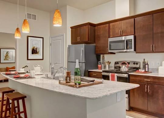 A modern kitchen here at 4800 Excelsior featuring a large island with light stone countertops, an undermount sink, and seating for casual dining. Dark wood cabinets line the walls alongside stainless steel appliances, while warm pendant lighting and coordinated décor connect the kitchen seamlessly to the adjacent living and dining area.