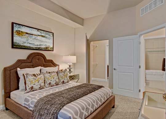 A serene bedroom here at 4800 Excelsior featuring a plush upholstered bed with patterned bedding, layered accent pillows, and a textured throw. Soft carpeting and neutral walls create a calming atmosphere, complemented by a bedside lamp, framed artwork, and a mirrored nightstand, with easy access to a walk-in closet and adjoining bathroom.