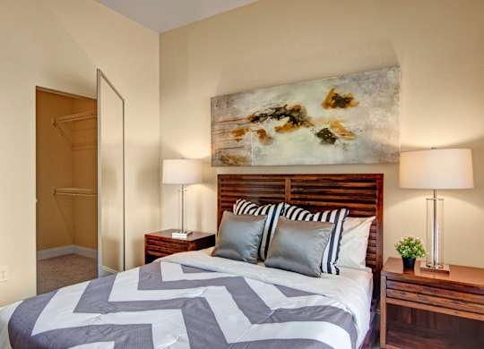A comfortable bedroom here at 4800 Excelsior featuring a wood bed frame with crisp patterned bedding and layered accent pillows. Matching nightstands with table lamps flank the bed beneath a large abstract art piece, while soft carpeting and neutral walls enhance the calm atmosphere, with a walk-in closet visible just off the sleeping area.