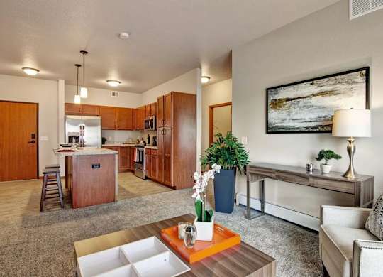 A living room here at Club at Heritage featuring an open layout with a warm-toned kitchen, wood cabinetry, stainless steel appliances, and a cozy seating area with a neutral armchair, console table, and decorative greenery.