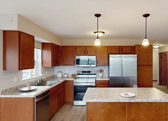 A kitchen here at Club at Heritage featuring rich wood cabinetry, stainless steel appliances, and a large island with pendant lighting, offering a bright and open layout that flows into the adjacent living space.