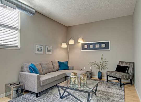 A cozy living room here at Marbella on Dean featuring a light gray sofa with blue accent pillows, a glass coffee table centered on a soft area rug, and neutral walls complemented by framed artwork, with an upholstered accent chair, modern floor lamp, exposed ductwork, and a window with blinds bringing in natural light.