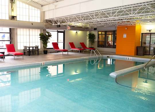 A bright indoor pool here at Marbella on Dean featuring a rectangular swimming pool with clear blue water, stainless steel handrails, and a wide concrete deck lined with red cushioned lounge chairs, accented by potted greenery, large frosted windows, and an industrial-style ceiling with exposed beams that allow soft natural light to fill the space.