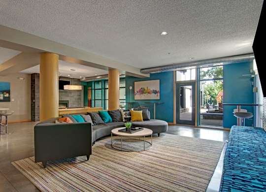 A modern community lounge here at Marbella on Dean featuring a large sectional sofa with colorful accent pillows arranged on a striped area rug, round coffee tables, and warm wood columns, with teal accent walls, contemporary artwork, high-top seating, and floor-to-ceiling windows and glass doors that bring in natural light and connect to an outdoor patio.
