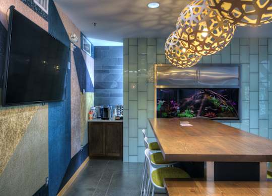 A modern community gathering space here at Marbella on Dean featuring a long wooden table with contemporary seating beneath sculptural pendant lighting, accented by textured walls, a large wall-mounted television, and a built-in aquarium centerpiece, with polished concrete floors and adjacent refreshment cabinetry creating a stylish and functional social setting.