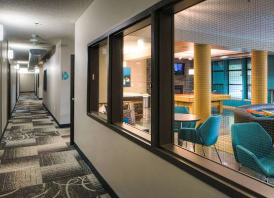 A welcoming interior hallway here at Marbella on Dean featuring patterned carpet flooring, neutral walls, and modern wall lighting, with large interior windows offering views into a vibrant community lounge furnished with teal seating, wood columns, and recreational tables, creating a connected and inviting atmosphere between shared spaces.