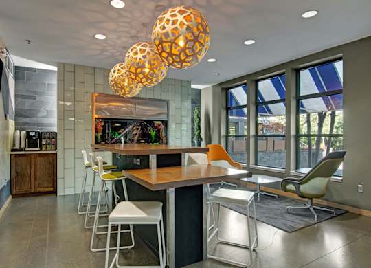 A stylish community gathering space here at Marbella on Dean featuring a long wood communal table with modern bar seating, sculptural pendant lighting overhead, and polished concrete flooring, complemented by a built-in aquarium wall, large windows with natural light, and cozy lounge chairs that create a social and inviting atmosphere.