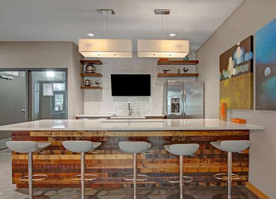 A modern community kitchen here at Marbella on Dean showcasing a long wood-front island with bar seating, smooth white countertops, and contemporary pendant lighting above, complemented by open wood shelving, a sleek backsplash, stainless-steel appliances, and colorful abstract artwork that creates a polished and social gathering space.