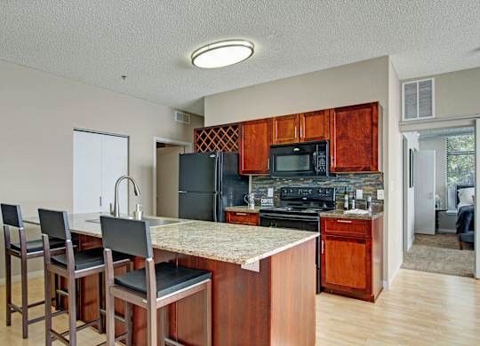 A warm open kitchen here at Marbella on Dean featuring rich wood cabinetry, a spacious island with granite-style countertops and bar seating, a tile backsplash, and a full appliance suite, with light wood flooring, a built-in wine rack, and an adjacent hallway leading toward the bedroom area creating a functional and inviting layout.