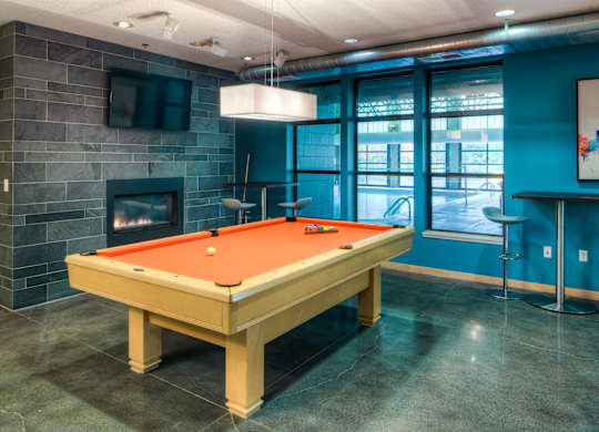 A stylish community game room here at Marbella on Dean featuring a full-size pool table with vibrant felt centered on polished concrete floors, surrounded by teal accent walls, a modern stone fireplace with mounted TV, pendant lighting, and large interior windows that look into the indoor pool area for an energetic and social atmosphere.
