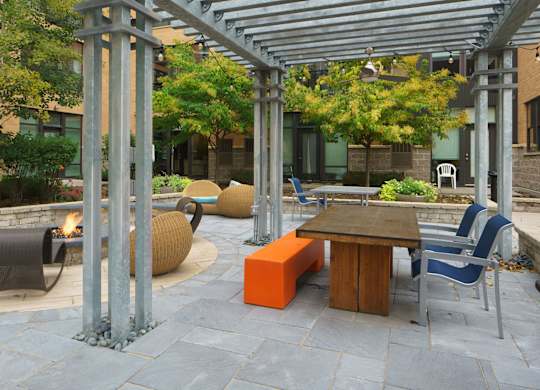 An inviting outdoor courtyard here at Marbella on Dean featuring a covered pergola with metal beams, string lights, and ceiling fans, set over stone paver flooring with a wooden communal table, an orange bench, and modern seating, alongside a circular fire pit, woven lounge chairs, raised planters, and lush trees surrounded by the apartment buildings.