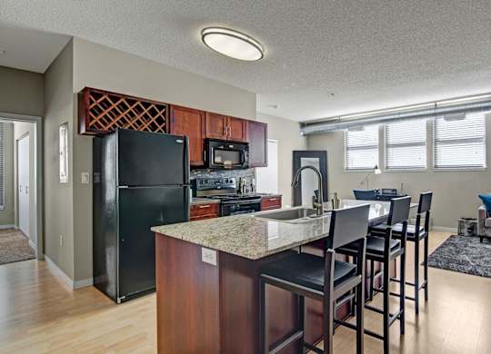 A spacious open kitchen here at Marbella on Dean showcasing rich wood cabinetry, a large island with granite-style countertops and bar seating, a tile backsplash, and a full appliance suite, flowing into the dining and living area with light wood flooring, exposed ductwork, and wide windows that bring in natural light.