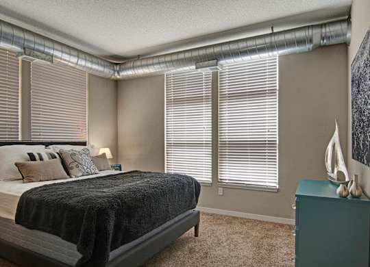 A bright industrial-inspired bedroom here at Marbella on Dean featuring a comfortable bed with layered neutral bedding and accent pillows, exposed metal ductwork along the ceiling, large windows with blinds that bring in natural light, soft carpeted flooring, and a teal dresser with modern decor creating a stylish and inviting retreat.