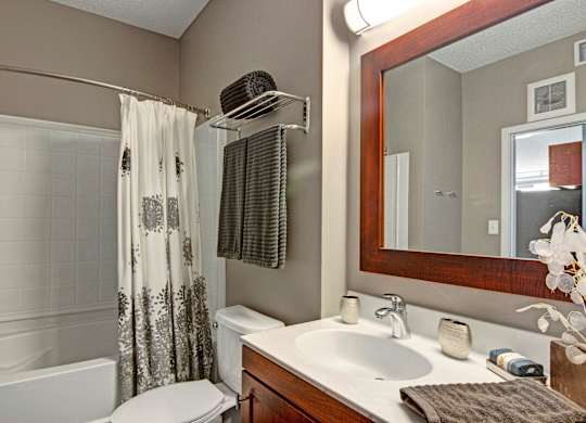 A contemporary bathroom here at Marbella on Dean featuring a white vanity with warm wood cabinetry, an integrated sink with chrome fixtures, a large framed mirror with overhead lighting, neatly folded towels on a wall-mounted rack, a shower with a patterned curtain, and soft neutral walls that create a clean, comfortable atmosphere.