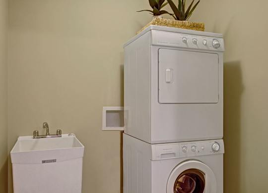 A convenient laundry area here at The Mist featuring a stacked washer and dryer set positioned beside a deep utility sink with a chrome faucet. Neutral wall tones create a clean look, while the compact layout maximizes functionality and provides an efficient space for everyday laundry tasks within the apartment.